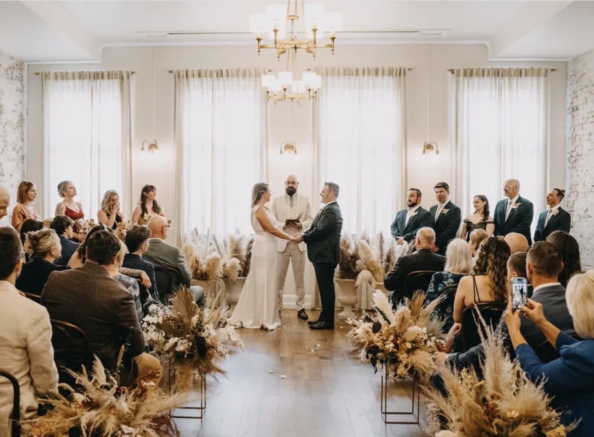 A wedding ceremony in a light-filled hall, guests seated among tall dried florals