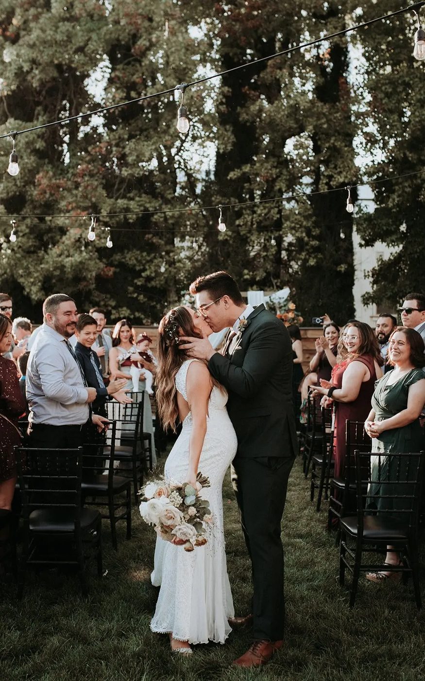 A couple kissing in the aisle after their outdoor ceremony, guests applauding under string lights